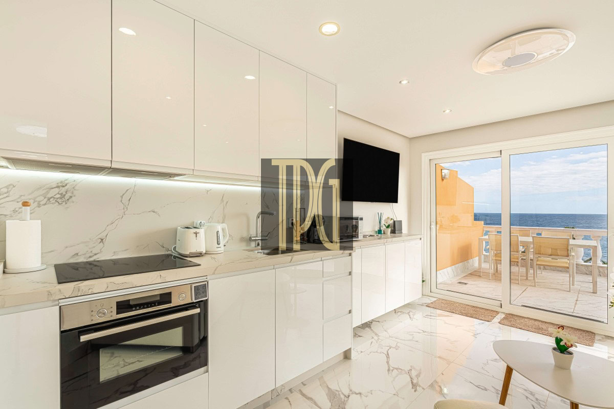 Bright white kitchen with marble countertops and cabinets, built-in oven, and a balcony door showing the ocean view.