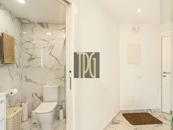 Modern white bathroom with marble walls and floor, toilet, towel on rack, and a glass shower door at left.