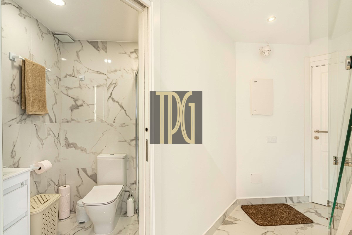 Modern marble-tiled bathroom with a white toilet, towel rack, laundry basket, and vanity on the left; sliding glass shower door hints at a separate shower area.