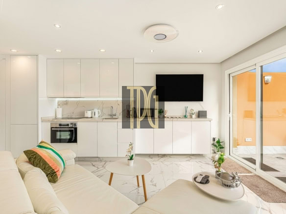 Bright open-plan living area with white glossy kitchen cabinets, marble floors, and a wall-mounted TV; sliding glass door to a sunny orange patio.