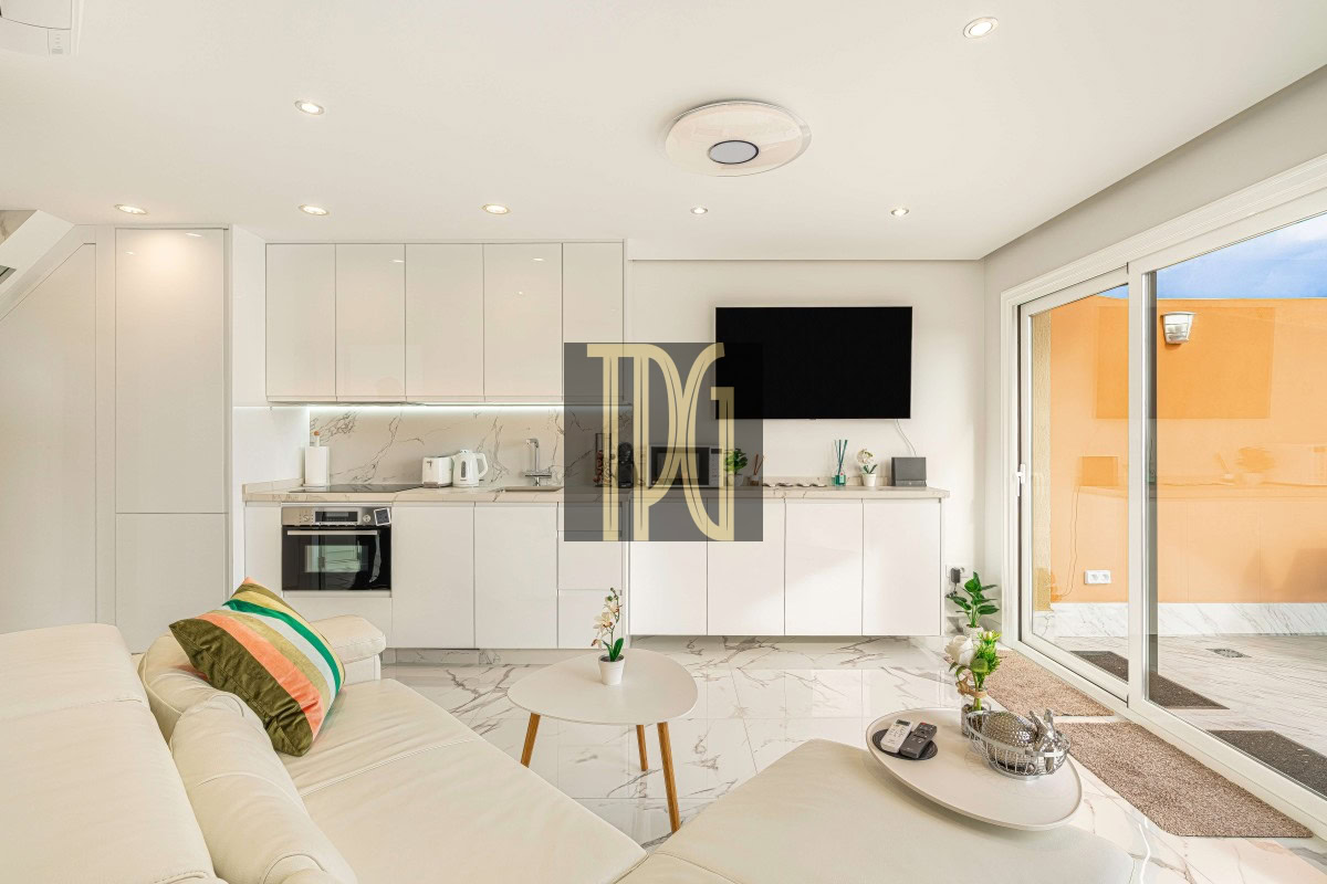 Bright open-plan living area with white glossy kitchen cabinets, marble floors, and a wall-mounted TV; sliding glass door to a sunny orange patio.