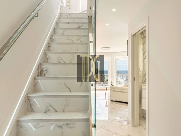Marble staircase with a glass railing beside a bright open hallway leading to a living area with a sea view through large windows.