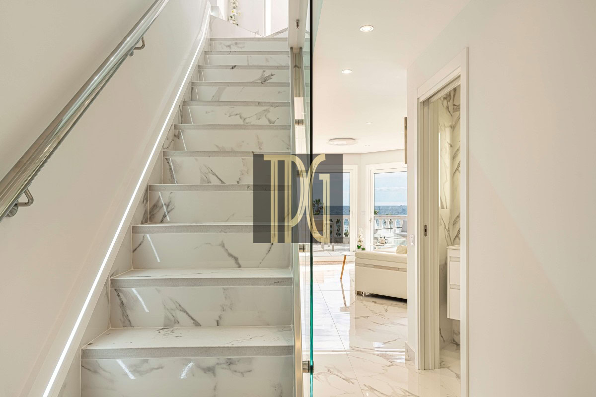 Marble staircase with a glass railing beside a bright open hallway leading to a living area with a sea view through large windows.