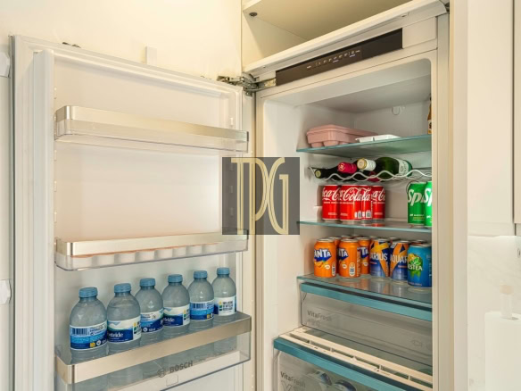 Open refrigerator showing water bottles on the left door shelves and a variety of cans and bottles (S-Cola, Fanta, Sprite, juices) on the right shelves in a clean, modern kitchen.