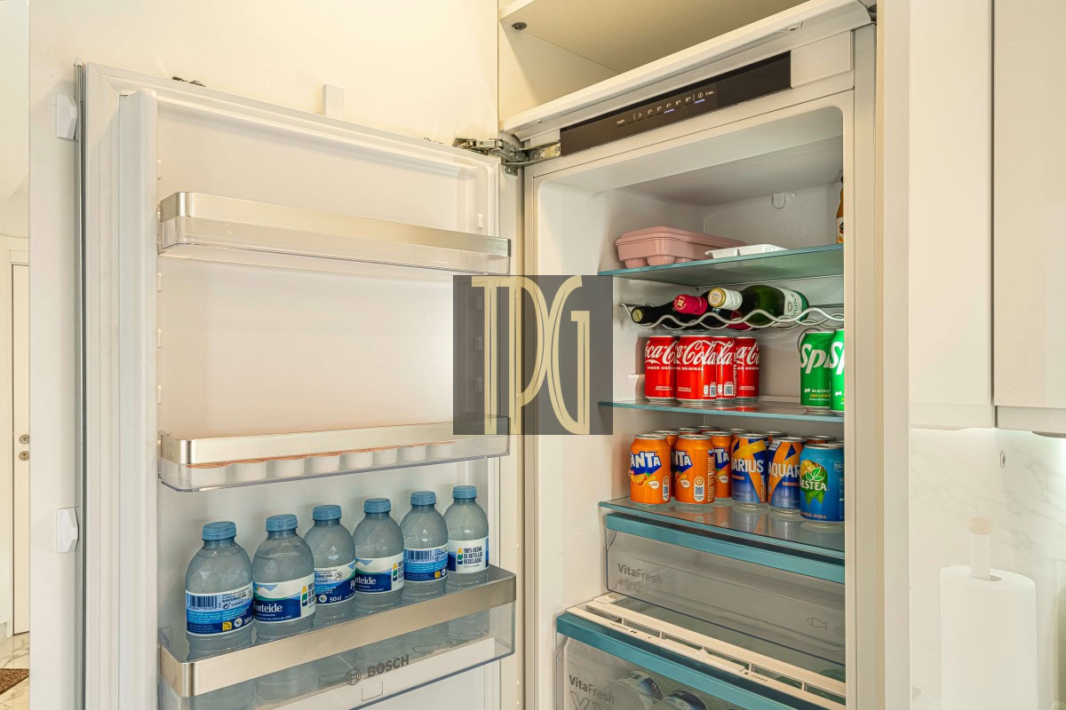 Open refrigerator showing water bottles on the left door shelves and a variety of cans and bottles (S-Cola, Fanta, Sprite, juices) on the right shelves in a clean, modern kitchen.