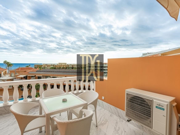 Balcony seating area with a glass-top table and four wicker chairs, an orange wall, and an ocean view beyond resort rooftops.