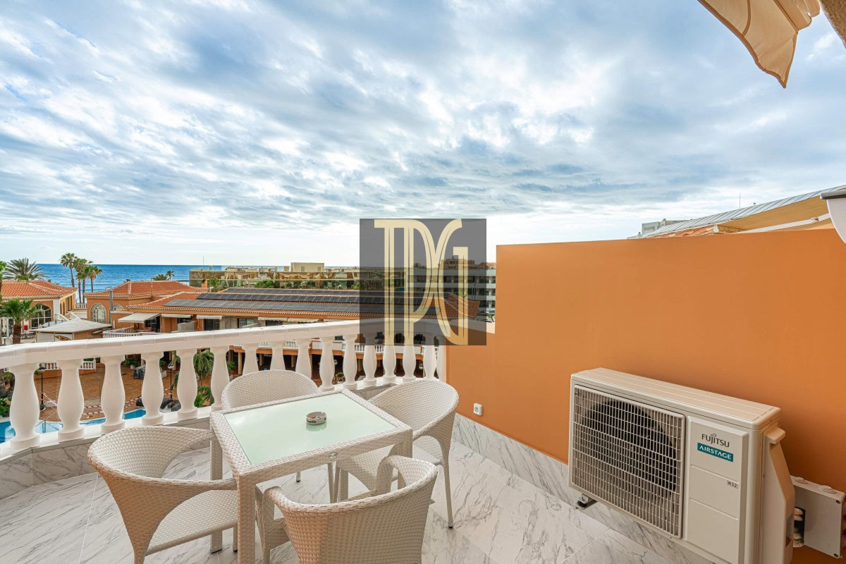 Balcony seating area with a glass-top table and four wicker chairs, an orange wall, and an ocean view beyond resort rooftops.