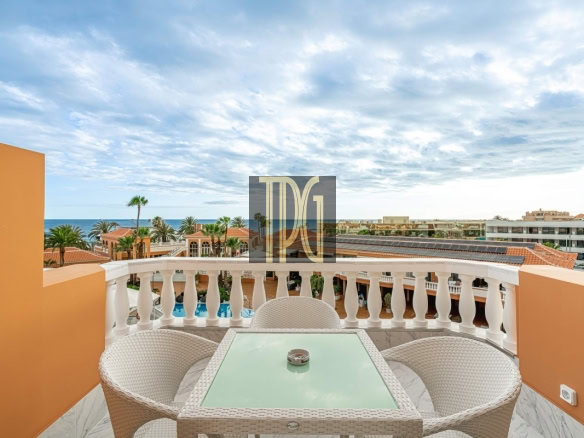 Balcony seating area with a glass-topped wicker table and white chairs, overlooking a resort pool, palm trees, and the ocean under a cloudy sky.