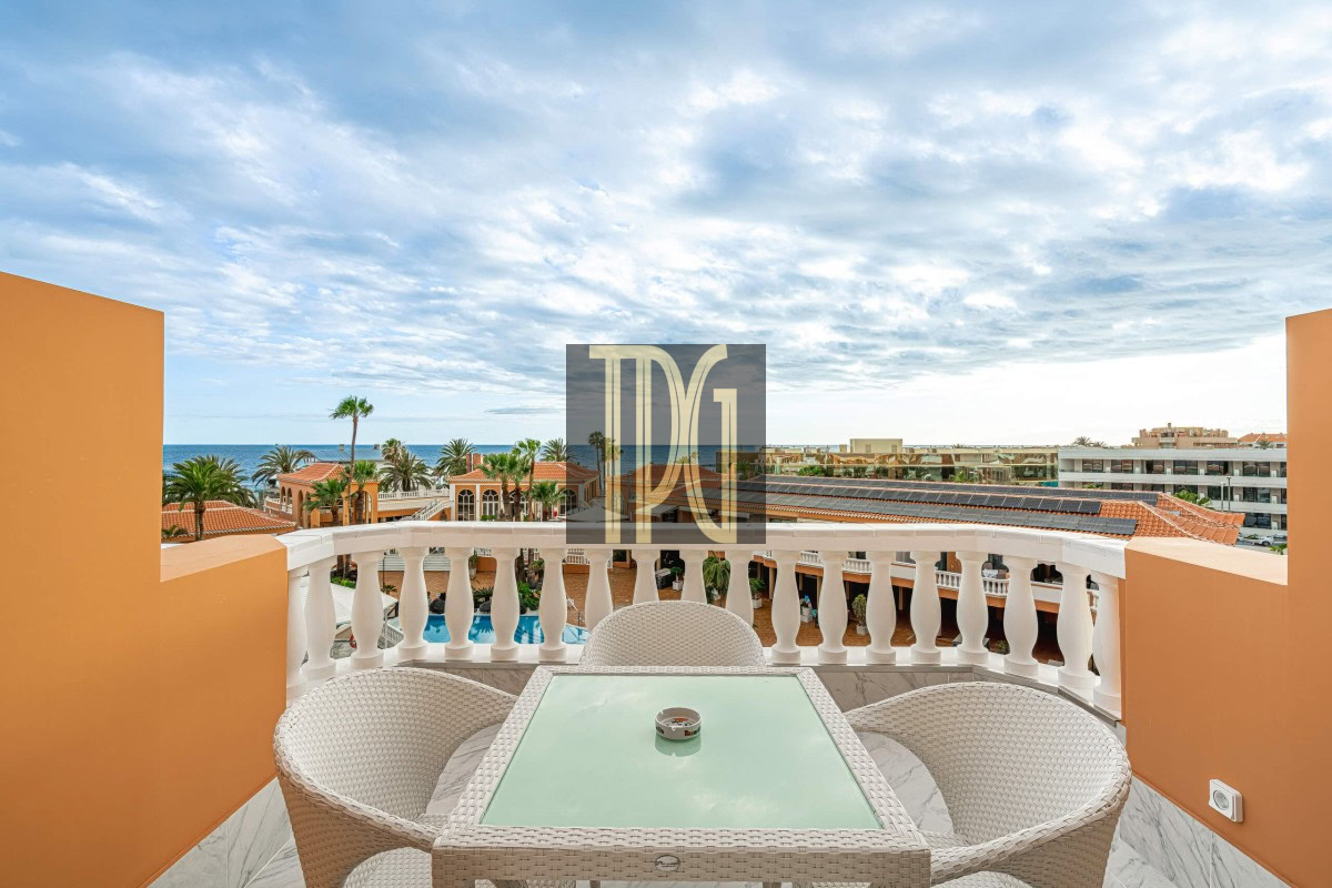 Balcony seating area with a glass-topped wicker table and white chairs, overlooking a resort pool, palm trees, and the ocean under a cloudy sky.