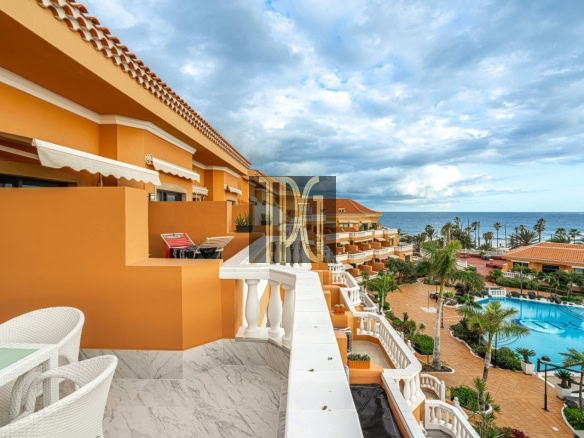 Terracotta-colored hotel complex with white railings and balconies, overlooking a large pool and the ocean beyond.