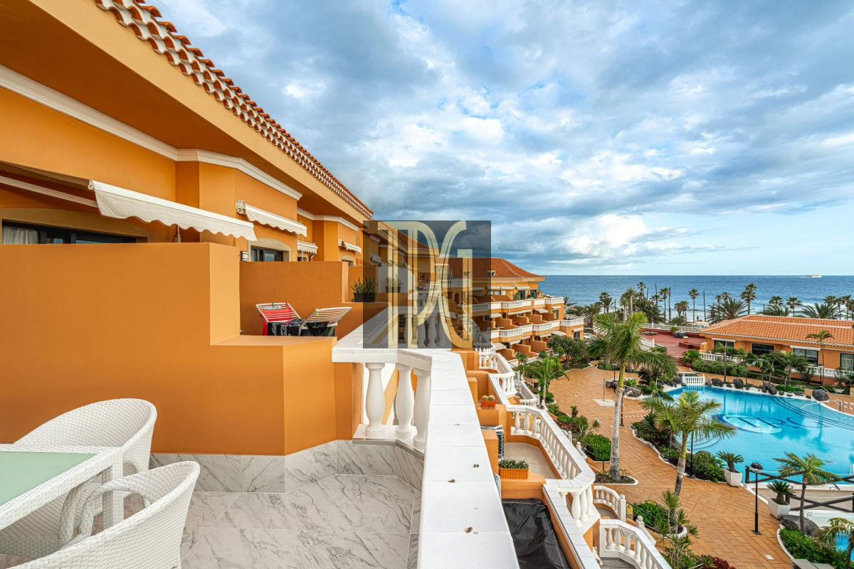 Terracotta-colored hotel complex with white railings and balconies, overlooking a large pool and the ocean beyond.