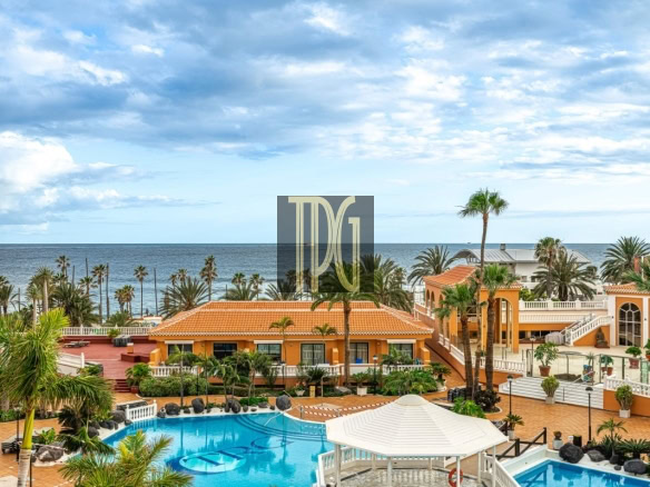 Resort courtyard with a blue swimming pool, orange-tiled buildings, and palm trees overlooking the ocean.