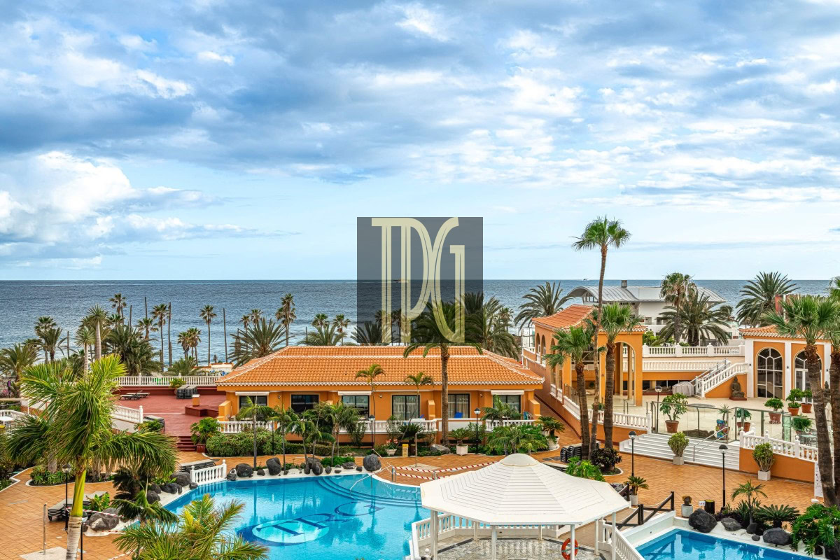 Resort courtyard with a blue swimming pool, orange-tiled buildings, and palm trees overlooking the ocean.