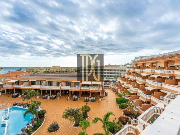 Resort courtyard with an orange-hued building and a large swimming pool, palm trees, and sun loungers facing the sea.