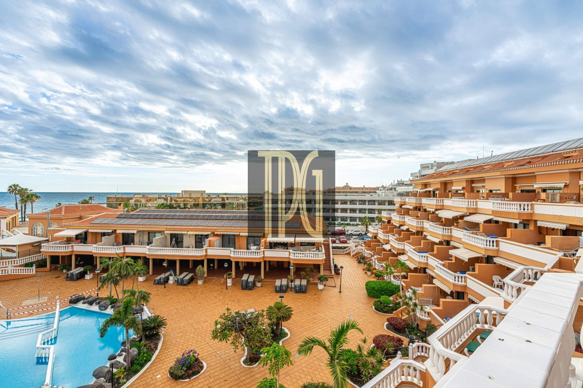 Resort courtyard with an orange-hued building and a large swimming pool, palm trees, and sun loungers facing the sea.