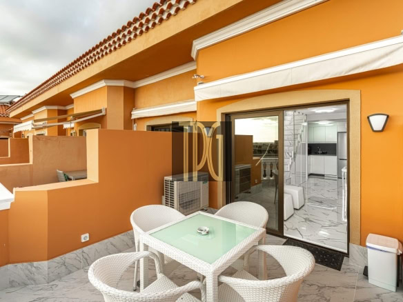 Orange-walled balcony with a white wicker dining set and glass-table, sliding doors opening to a modern kitchen inside.