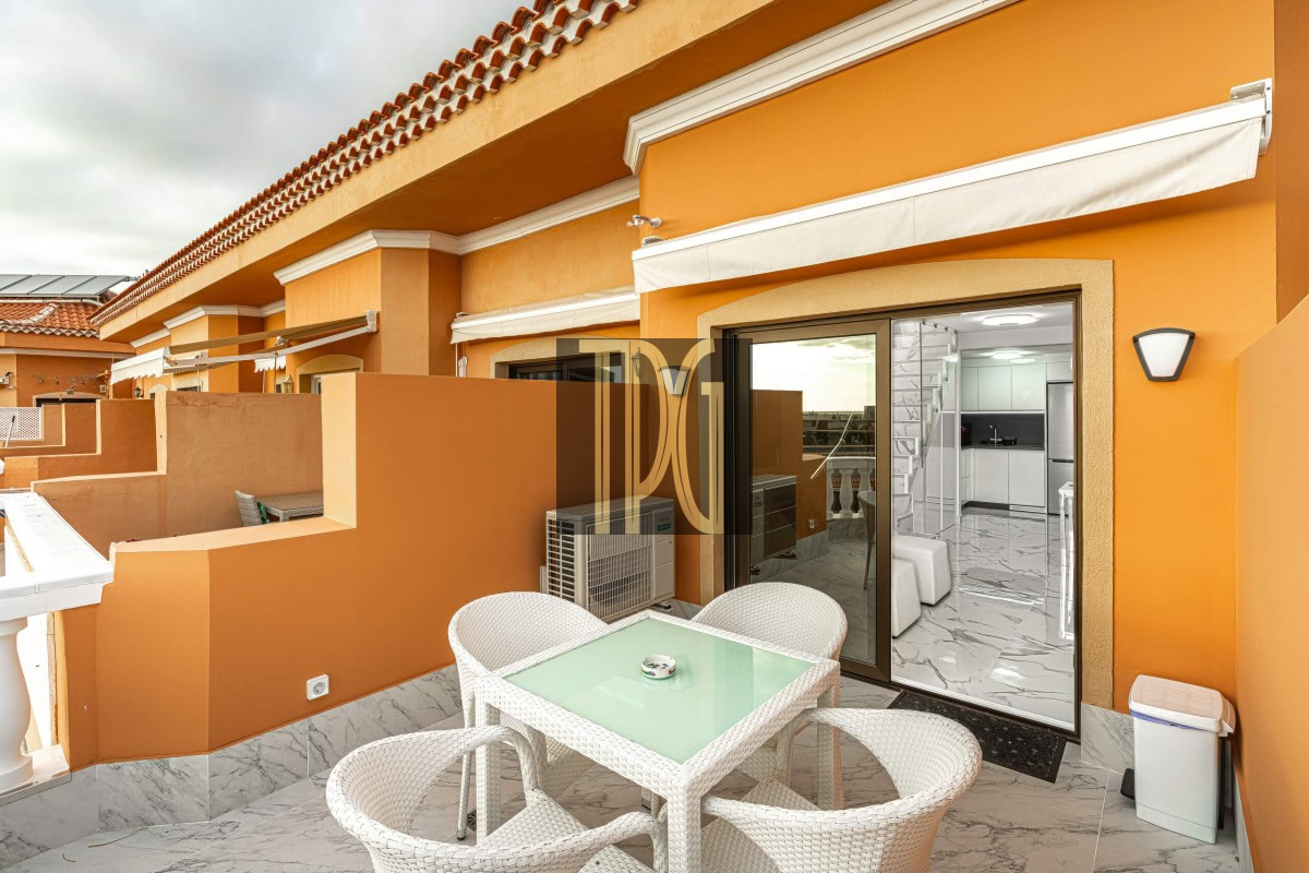Orange-walled balcony with a white wicker dining set and glass-table, sliding doors opening to a modern kitchen inside.