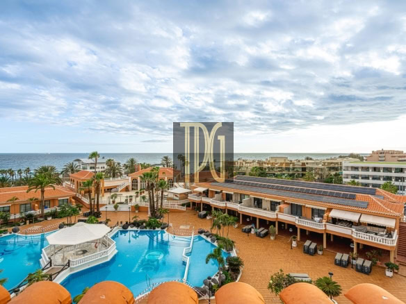 Resort pool area with blue water, orange-tiled patios, palm trees, and buildings overlooking the ocean.