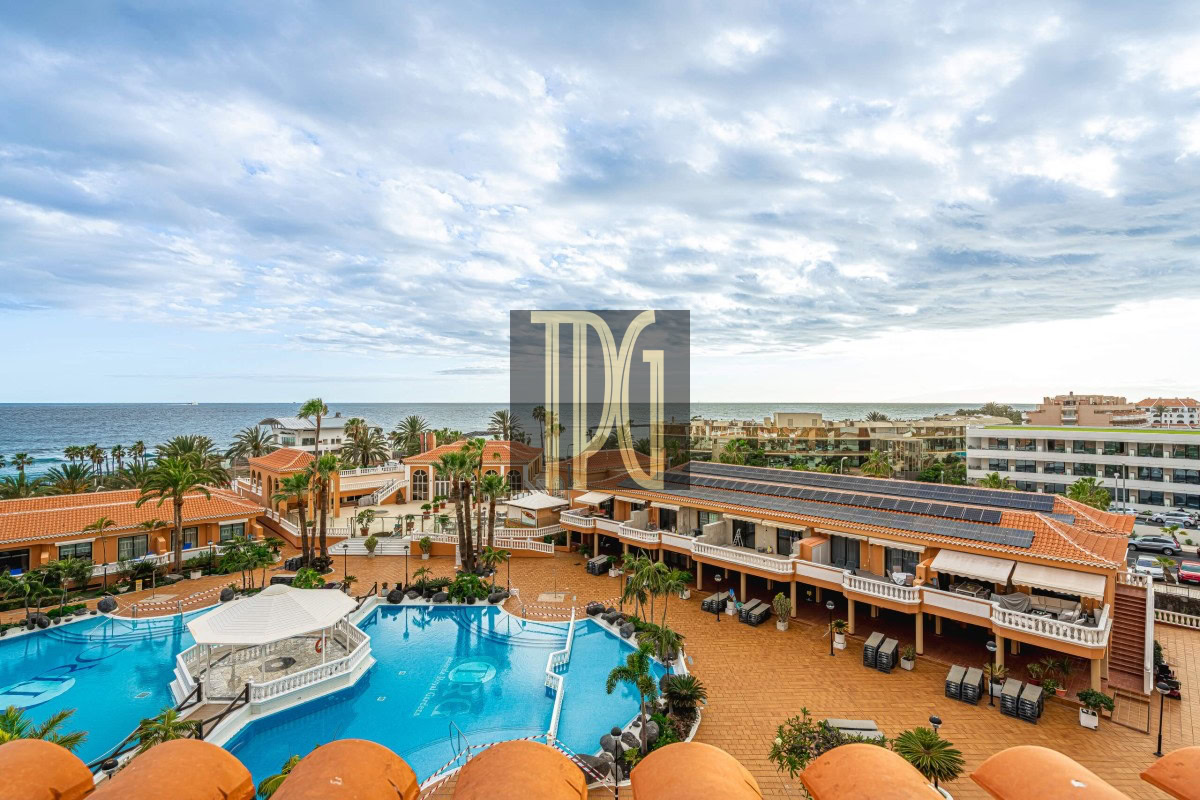 Resort pool area with blue water, orange-tiled patios, palm trees, and buildings overlooking the ocean.