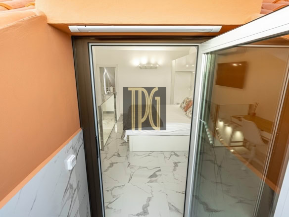View into a bright, modern bedroom with a white bed and marble floor seen from an orange-walled balcony through a sliding glass door, indoors lit warmly.