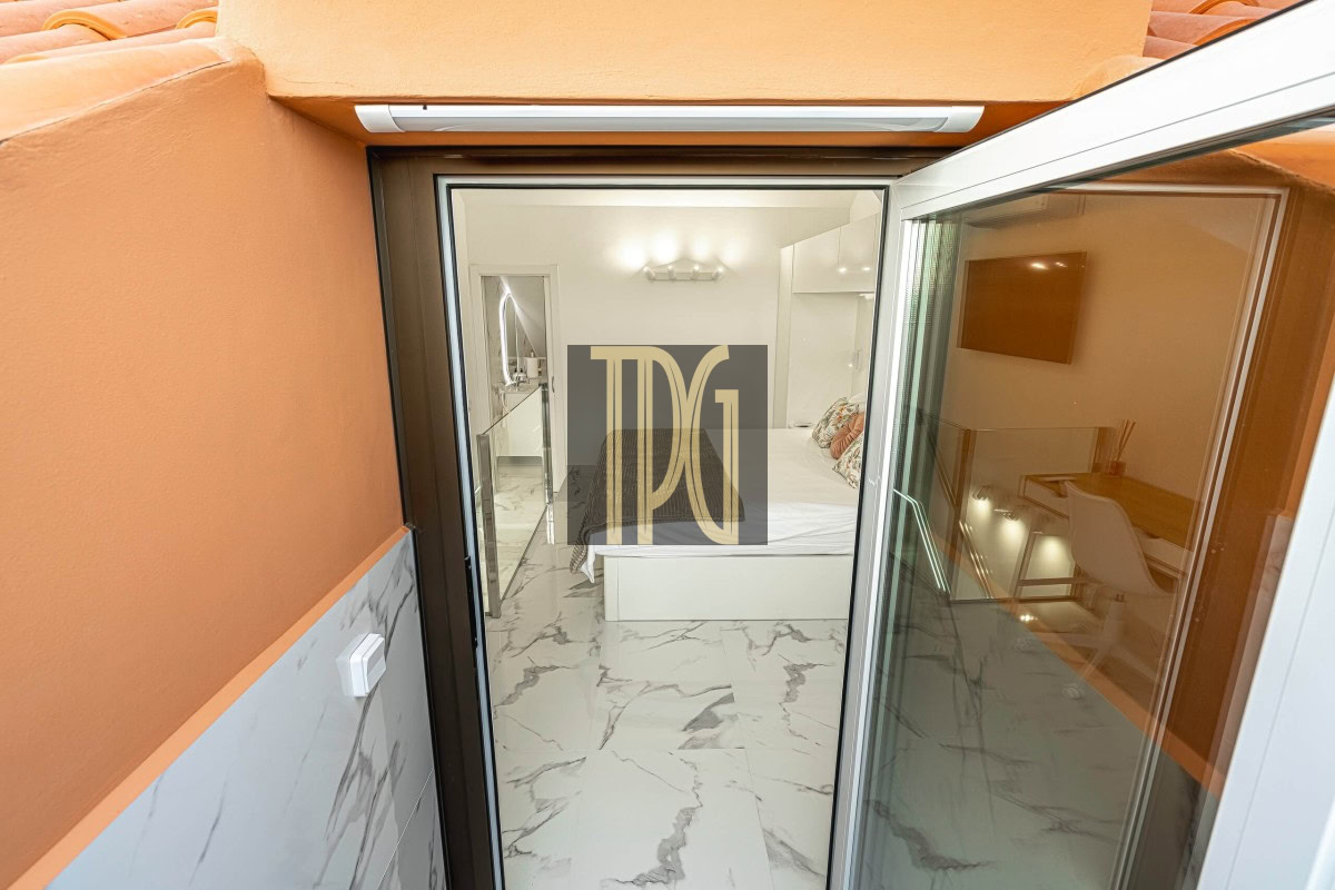 View into a bright, modern bedroom with a white bed and marble floor seen from an orange-walled balcony through a sliding glass door, indoors lit warmly.