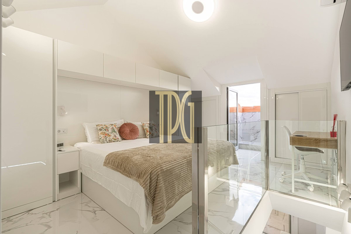 Bright white hotel bedroom with a king bed, beige throw, floral cushions, and a glass railing by the stairs.