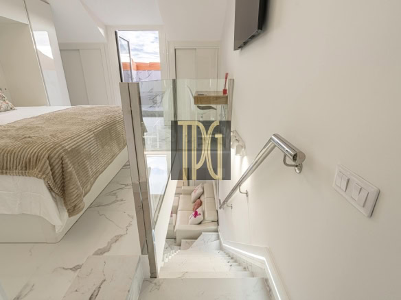 Modern bedroom with a raised bed, beige throw, floral pillow, white marble floor, and glass railing overlooking a lower lounge area.