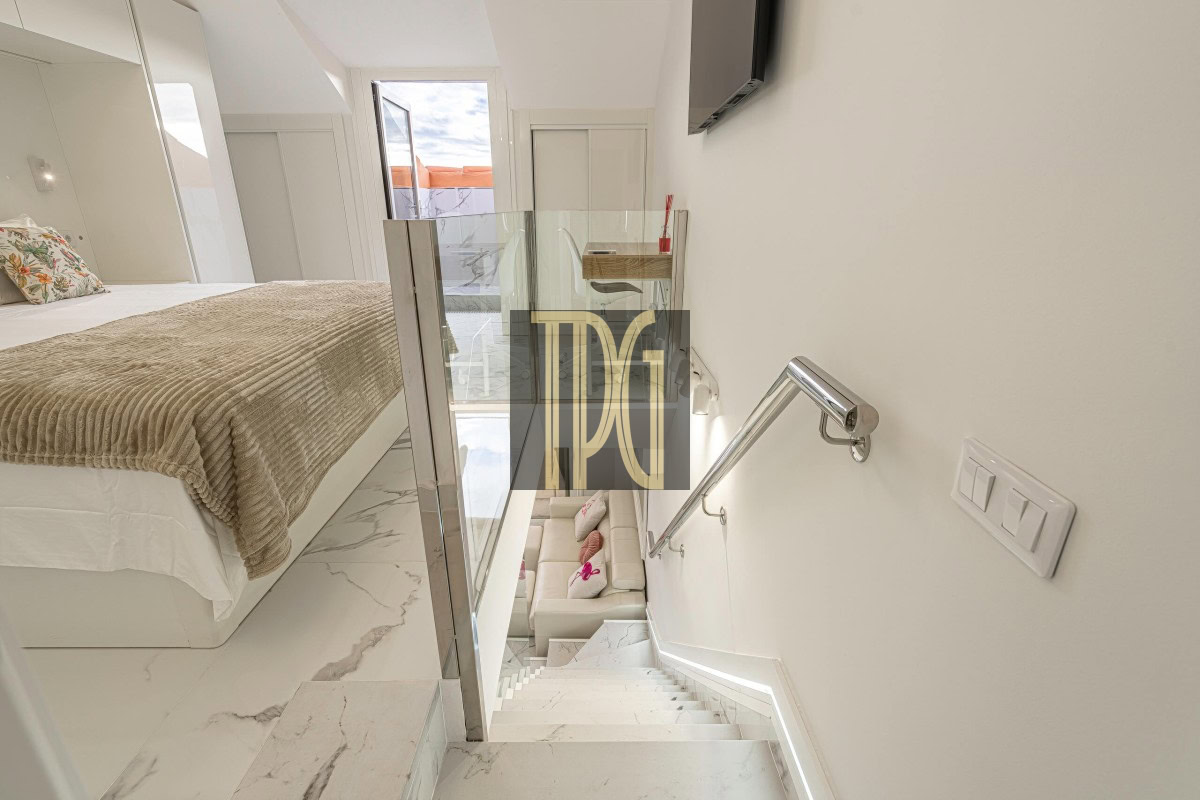 Modern bedroom with a raised bed, beige throw, floral pillow, white marble floor, and glass railing overlooking a lower lounge area.