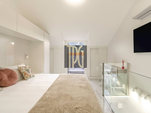 Bright white loft bedroom with a large bed, patterned pillows, and a beige textured throw running along the center.