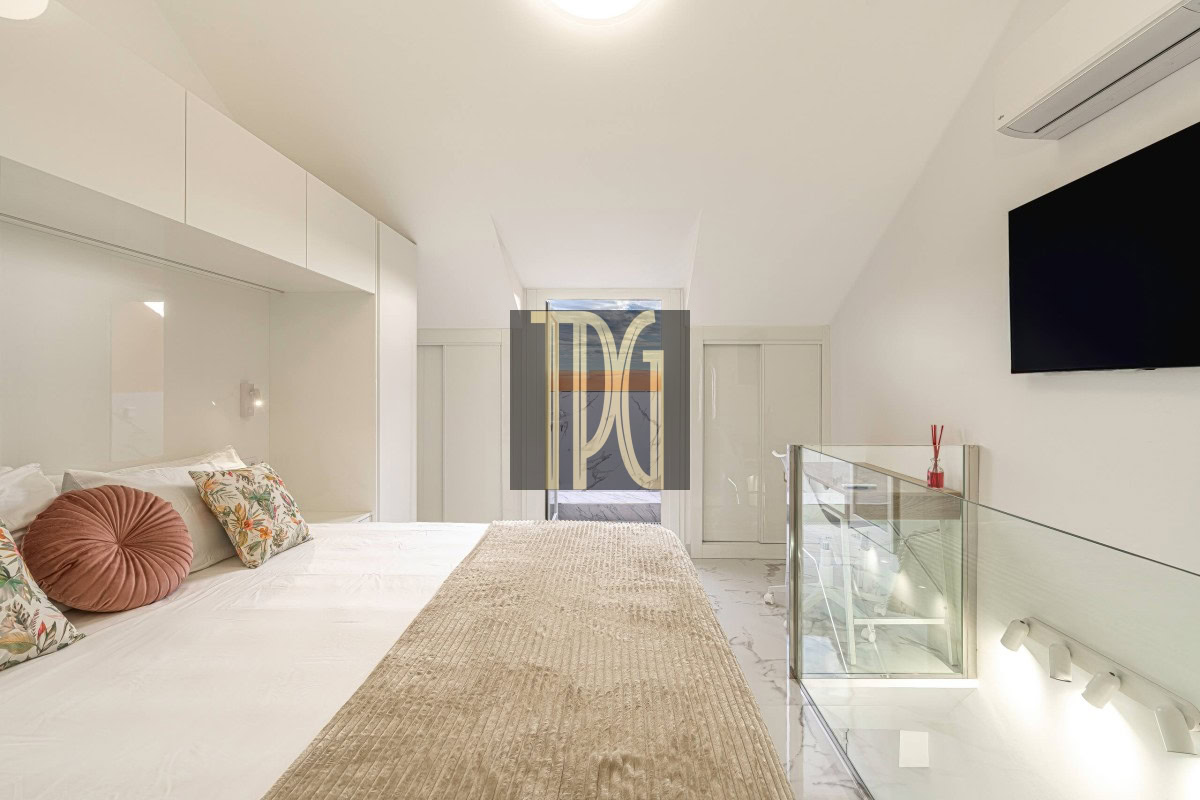 Bright white loft bedroom with a large bed, patterned pillows, and a beige textured throw running along the center.