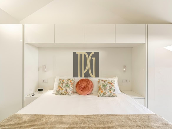 Minimalist white bedroom with built-in cabinetry surrounding a bed, featuring two floral cushions and a round coral pillow on top of white linens.”] ,