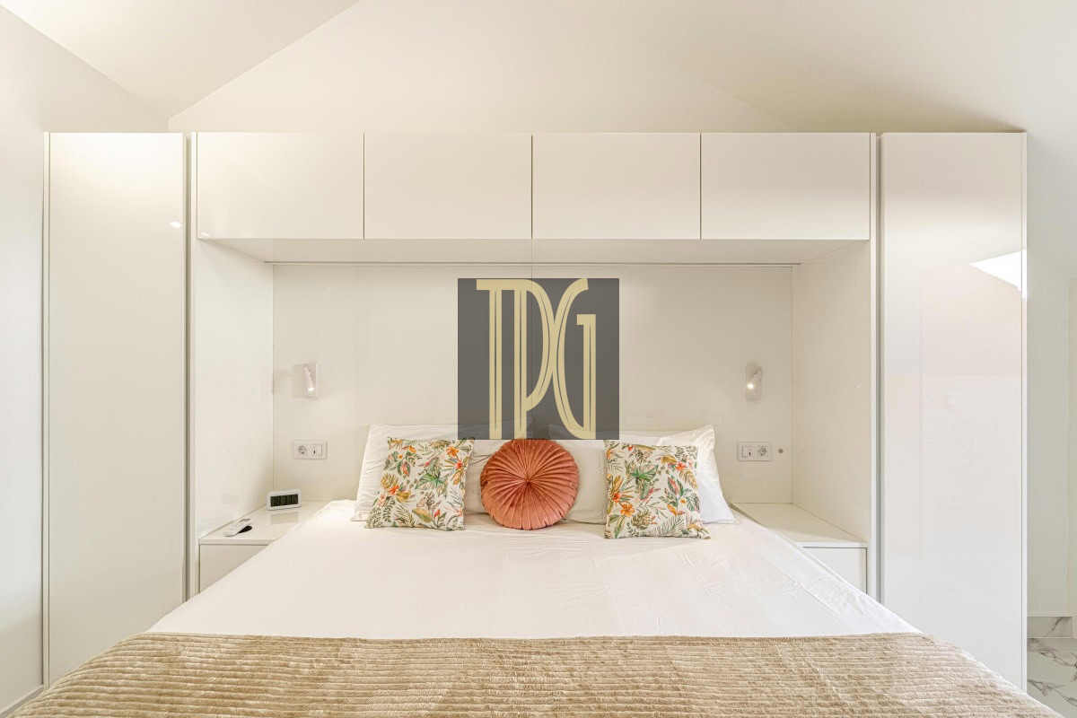 Minimalist white bedroom with built-in cabinetry surrounding a bed, featuring two floral cushions and a round coral pillow on top of white linens.”] ,