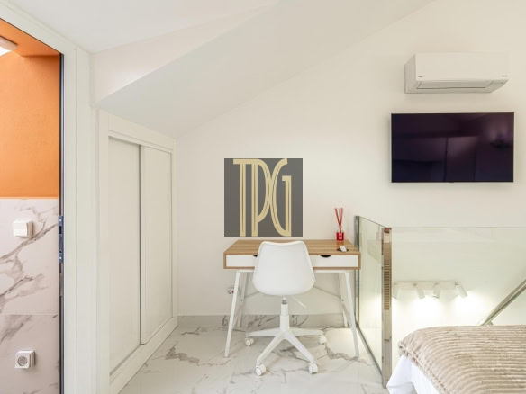 Modern loft bedroom with a wooden desk and white chair under a slanted ceiling, a wall-mounted TV, and marble floors.