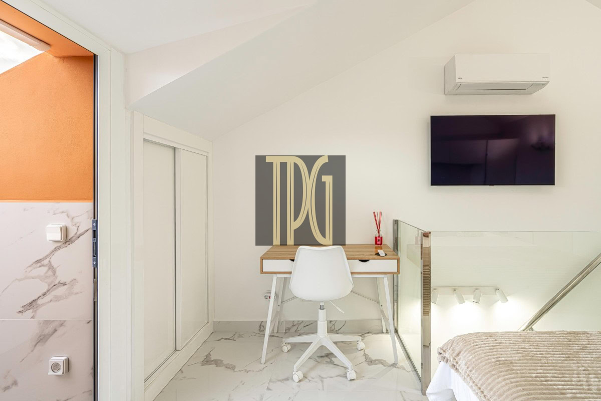 Modern loft bedroom with a wooden desk and white chair under a slanted ceiling, a wall-mounted TV, and marble floors.