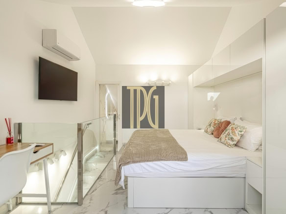 Bright modern bedroom with a white bed, decorative pillows, and a taupe throw; glass railing and marble floor create an open feel.