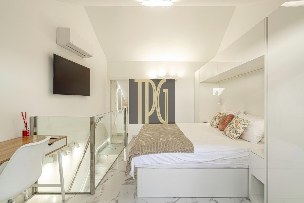 Bright modern bedroom with a white bed, decorative pillows, and a taupe throw; glass railing and marble floor create an open feel.