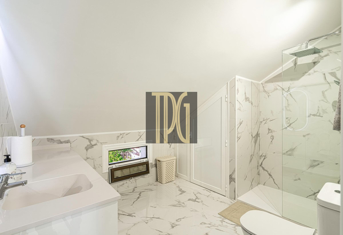 Bright marble bathroom with glass shower, white vanity, and small window.