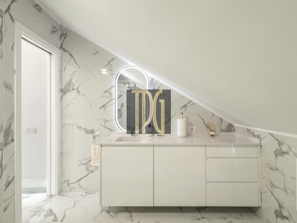 White bathroom with marble walls and floor, sloped ceiling, and a glossy vanity with sink and oval backlit mirror.