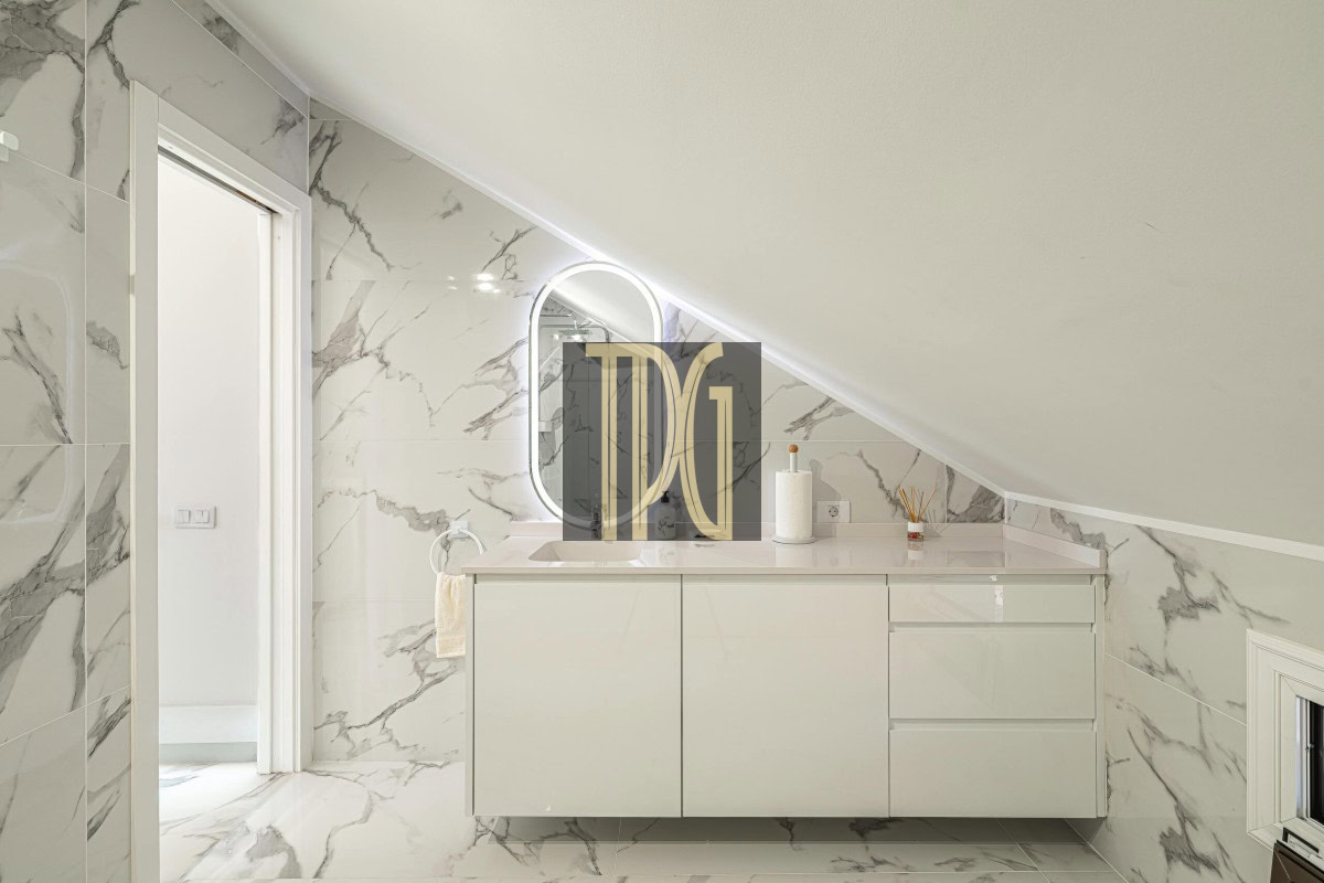 White bathroom with marble walls and floor, sloped ceiling, and a glossy vanity with sink and oval backlit mirror.