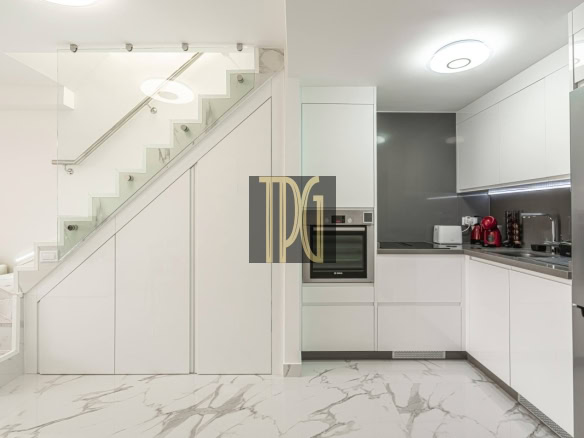 Modern white kitchen under a staircase with built-in oven, stainless fridge, and marble floors.