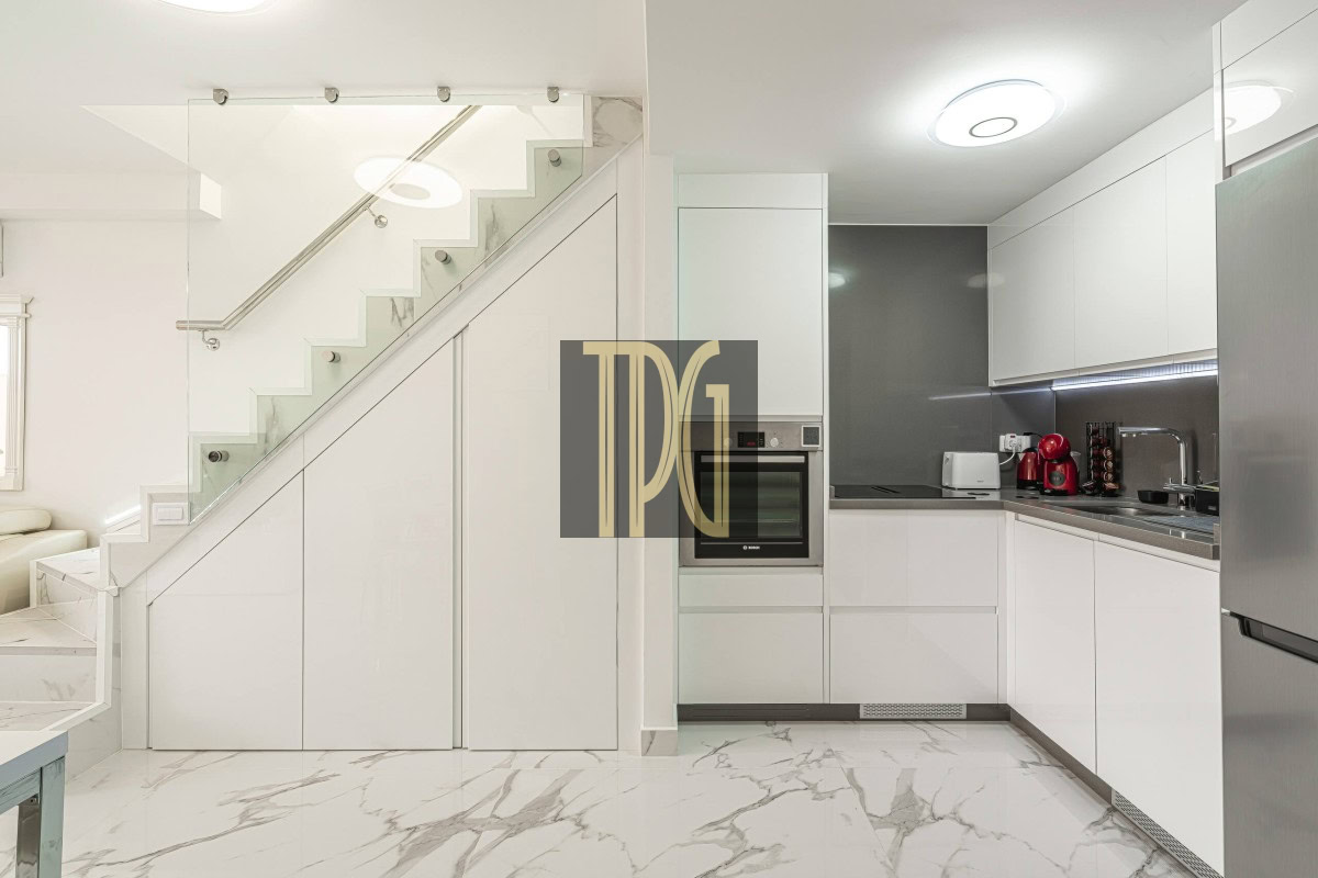 Modern white kitchen under a staircase with built-in oven, stainless fridge, and marble floors.