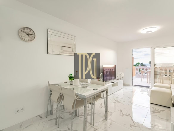 Bright open-plan living and dining area with white walls, marble floors, and a glass door to a balcony overlooking the ocean.