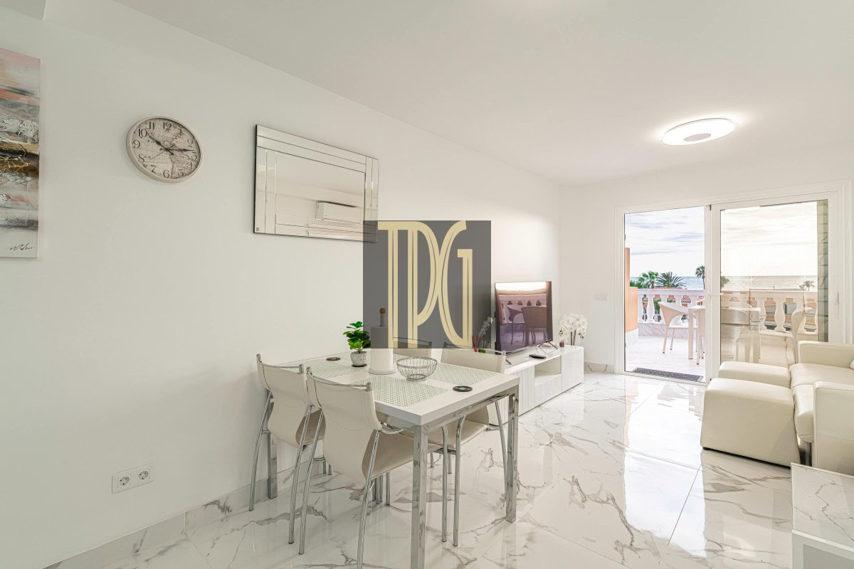 Bright open-plan living and dining area with white walls, marble floors, and a glass door to a balcony overlooking the ocean.