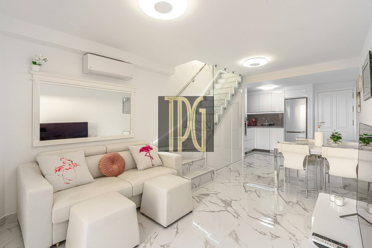 Bright open-concept living area with a white leather sofa, marble floors, glass-railed staircase, and a nearby dining/kitchen space.