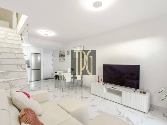 Bright open-plan living area with white sofa, marble floors, and a wall-mounted TV on a white media console; stairs with marble steps to the left.