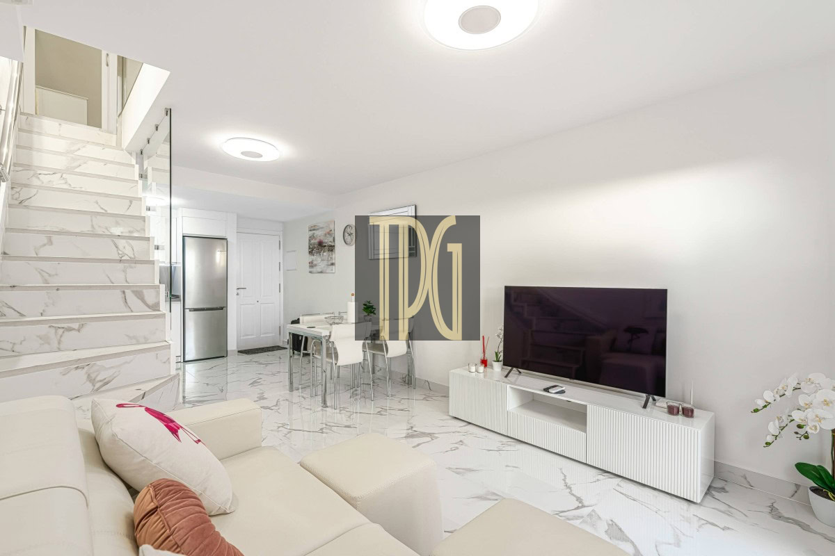 Bright open-plan living area with white sofa, marble floors, and a wall-mounted TV on a white media console; stairs with marble steps to the left.