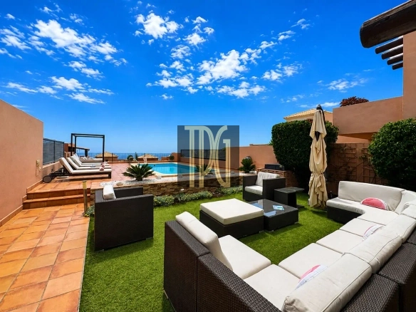 Sunlit rooftop patio with a rectangular pool, tan tiled deck, and white-cushioned sectional seating under a clear blue sky.