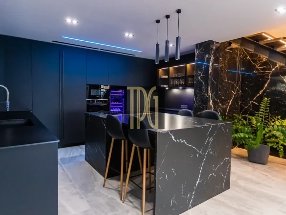 Modern dark kitchen with black marble island, three black stools, blue accent lighting, and potted plants.