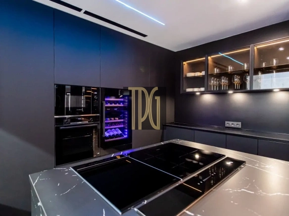 Dark modern kitchen with built-in ovens, a purple-lit wine fridge, glass-front cabinets, and a marble island with an induction cooktop.