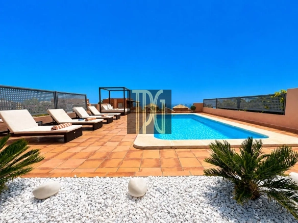 Rooftop pool area with blue water, brown tiled deck, white lounge chairs, and a metal railing against a clear blue sky